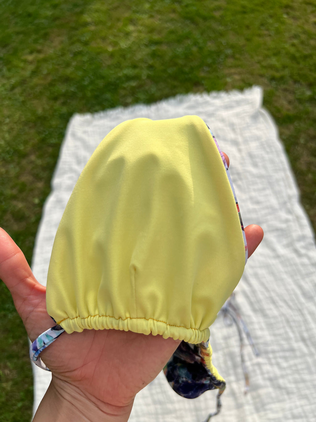 Hand holding a yellow bikini top with grass and a blanket in the background