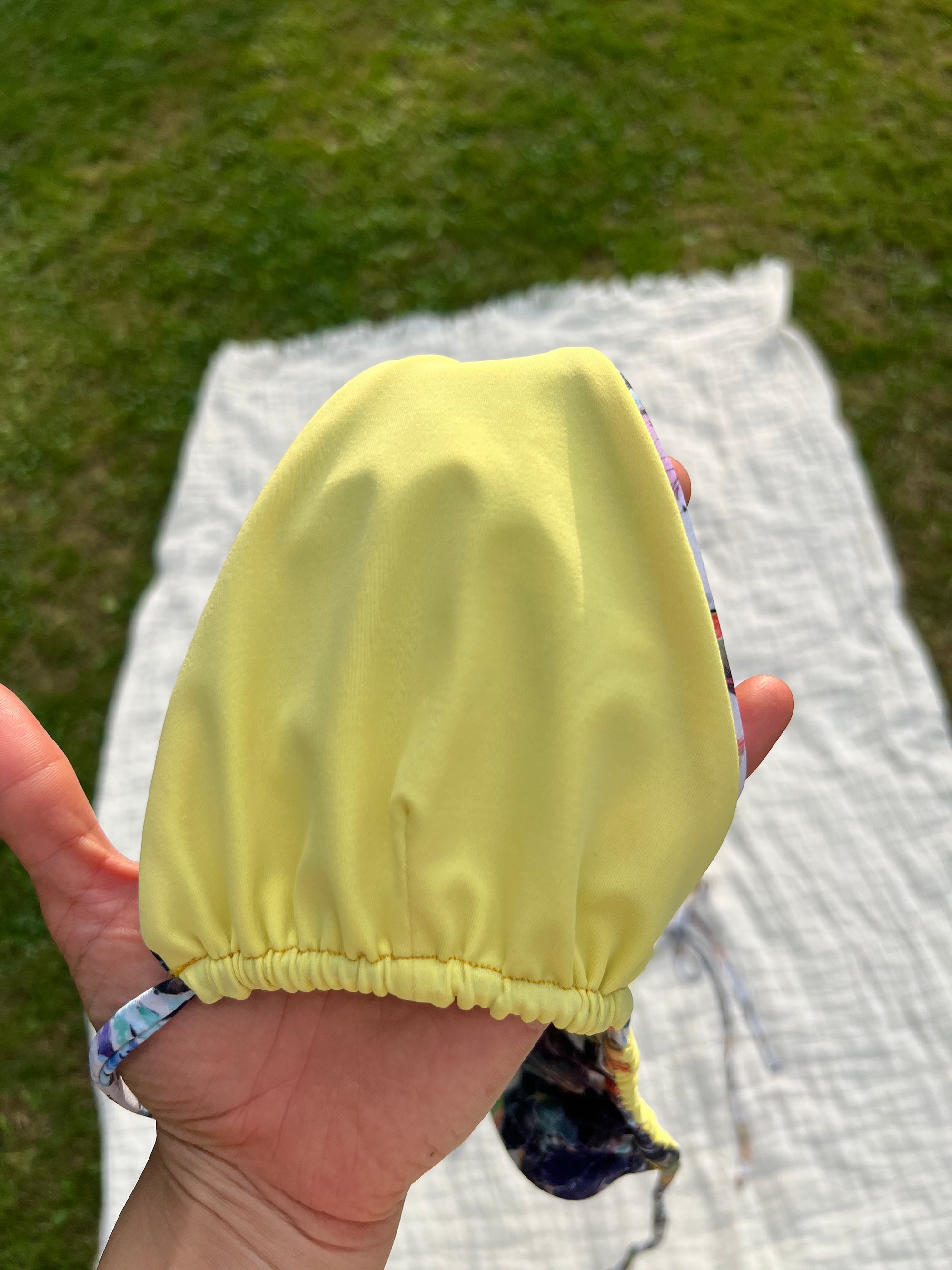 Hand holding a yellow bikini top with grass and a blanket in the background